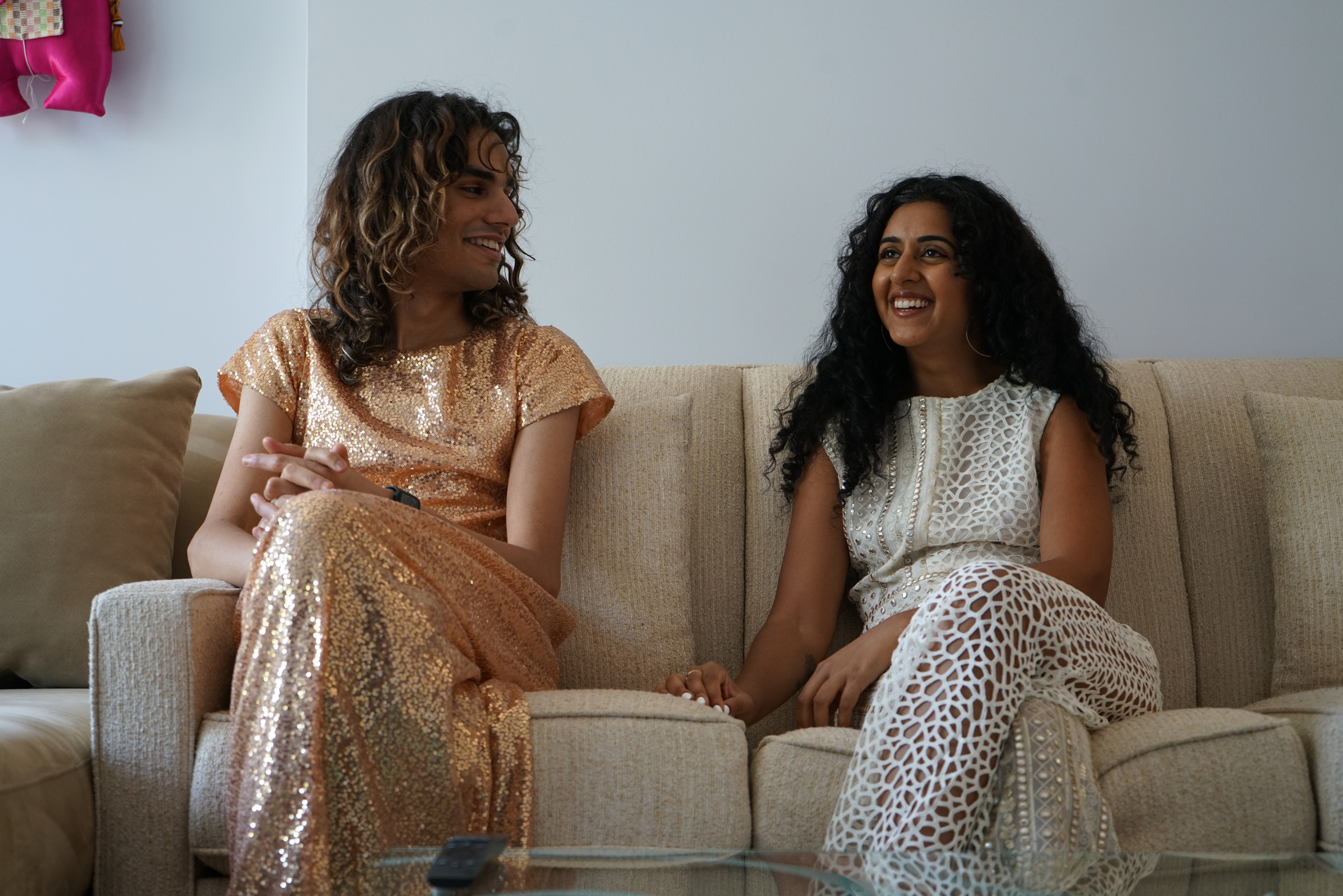 Sabrina and mayed sitting on a couch. mayed is a light brown person wearing a two piece gold sequins outfit, and has brown wavy hair with blonde highlights. sabrina is a medium brown person with black curly hair, wearing a white lace 2 piece outfit